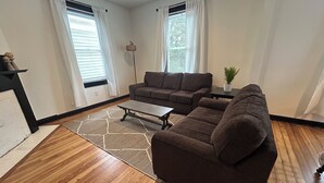 A living room with a brown sofa, a coffee table, and a rug. There are two windows with curtains, a floor lamp, and a small plant on a table. - The Uptown Suite - Big Red, 1903; Downtown; 2 bedroom 1 full bath, 3 queen beds (Wilmington)