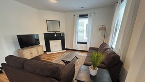 A living room with a sofa set, a TV, a fireplace, and a window with curtains. - The Uptown Suite - Big Red, 1903; Downtown; 2 bedroom 1 full bath, 3 queen beds (Wilmington)