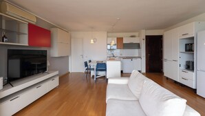 Ein modernes Wohnzimmer mit Flachbildfernseher, weißem Sofa, Essbereich mit Stühlen, Küche mit weißen Schränken und Holzboden.