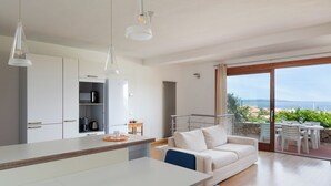 Eine moderne Küche mit weißen Schränken, ein Essbereich mit weißem Tisch und Stühlen sowie ein Balkon mit Meerblick.