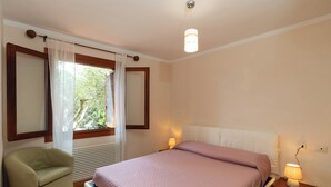 Ein Schlafzimmer mit einem Bett, einem Sessel, einem Nachttisch mit Lampe, einem Fenster mit Vorhängen und einer Deckenleuchte.
