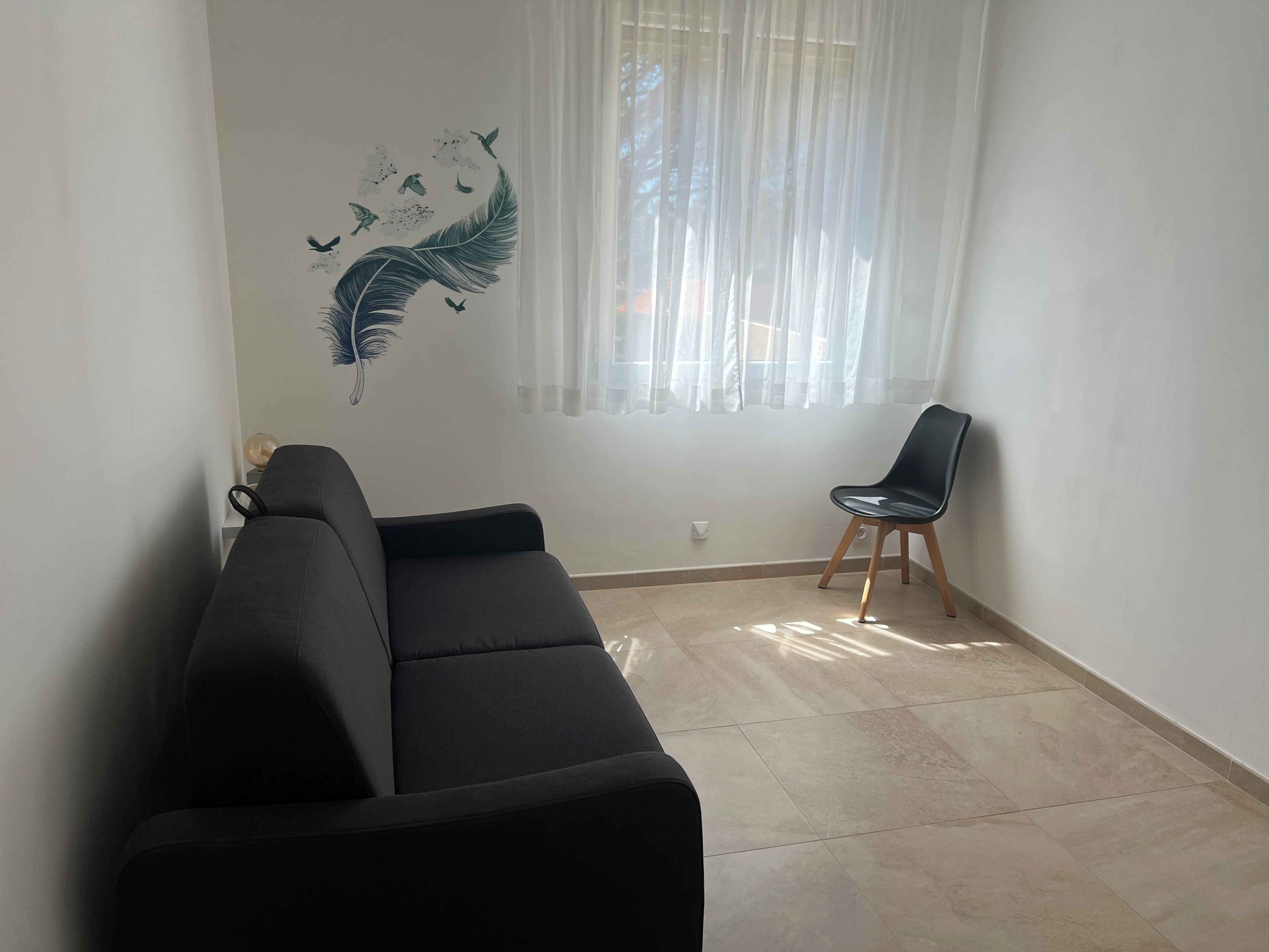 A living room with a black sofa, a wooden chair, a wall mural of birds, and a window with sheer curtains.