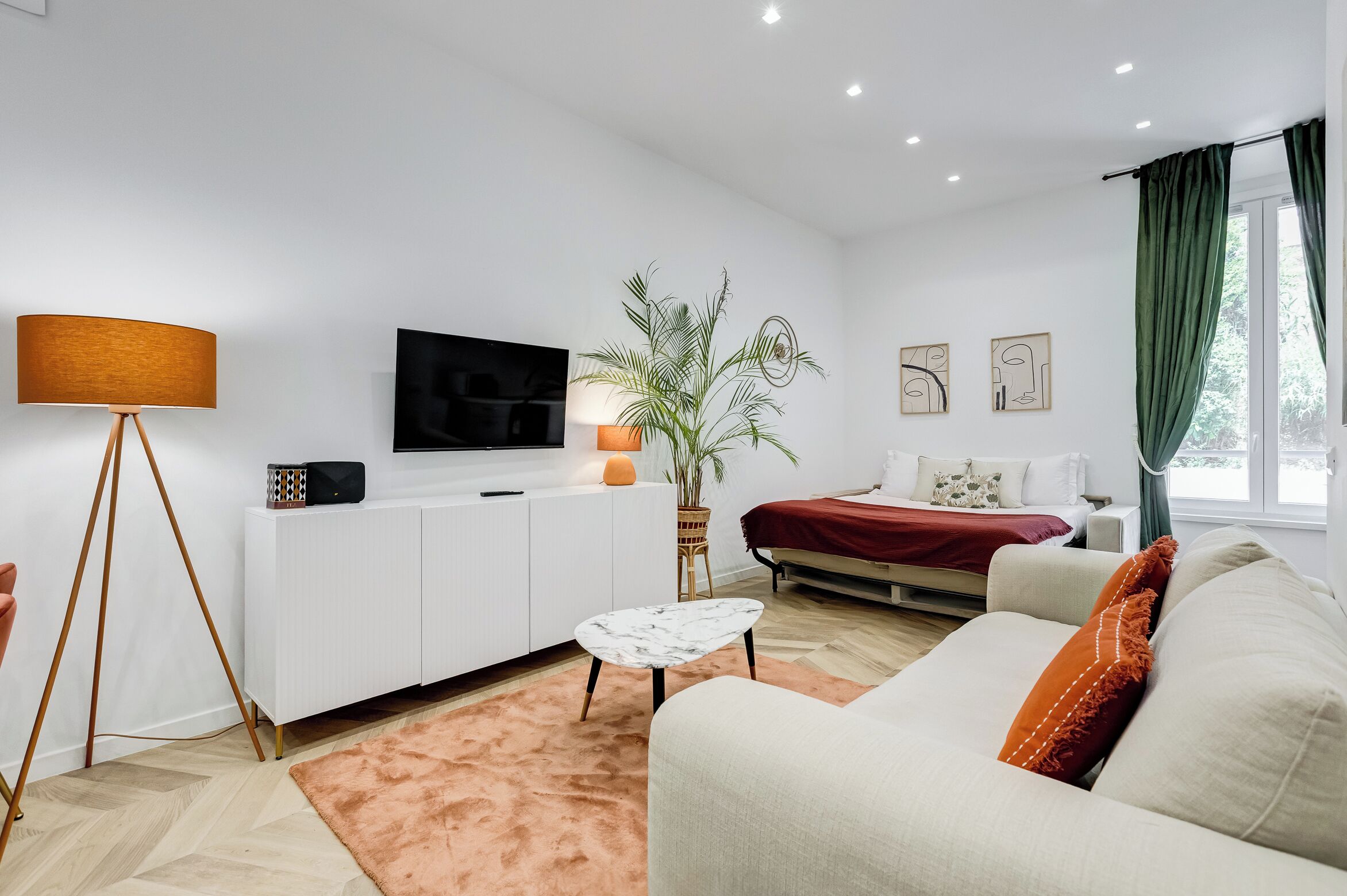 A modern living room with a sofa, a coffee table, a TV, a lamp, a plant, and a bed with a red blanket.