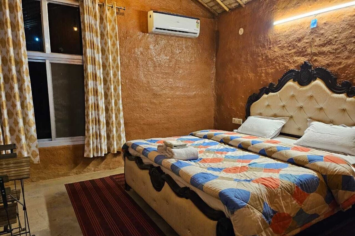 A double bed with a patterned quilt, a red rug, a window with curtains, and an air conditioning unit on the wall.