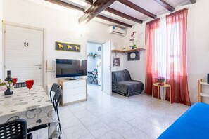 Ein möbliertes Zimmer mit Fernseher, Esstisch, Stühlen, einer Couch, einem kleinen Tisch mit Pflanze und einem Fenster mit Vorhängen.
