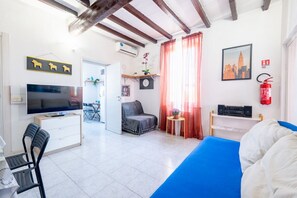 Ein Zimmer mit Bett, Fernseher, Schreibtisch, Stuhl, einer Uhr, einem Fenster mit Vorhängen, einem Feuerlöscher und einem Stadtbild.