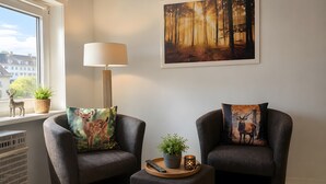 A cozy living room with two armchairs, a side table, a lamp, a fireplace, and a window with a view of buildings. - Apartment 'Rehkitz' with Mountain View (Bad Lauterberg im Harz)