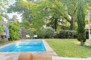 Ein rechteckiger Pool mit Steinrand, umgeben von einer Rasenfläche und verschiedenen Bäumen.