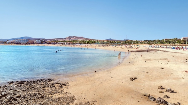 A beach with clear water, sandy shore, and buildings in the background.
