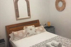 Een net opgemaakt bed met een houten hoofdbord, een nachtkastje met een lamp en een spiegel aan de muur.