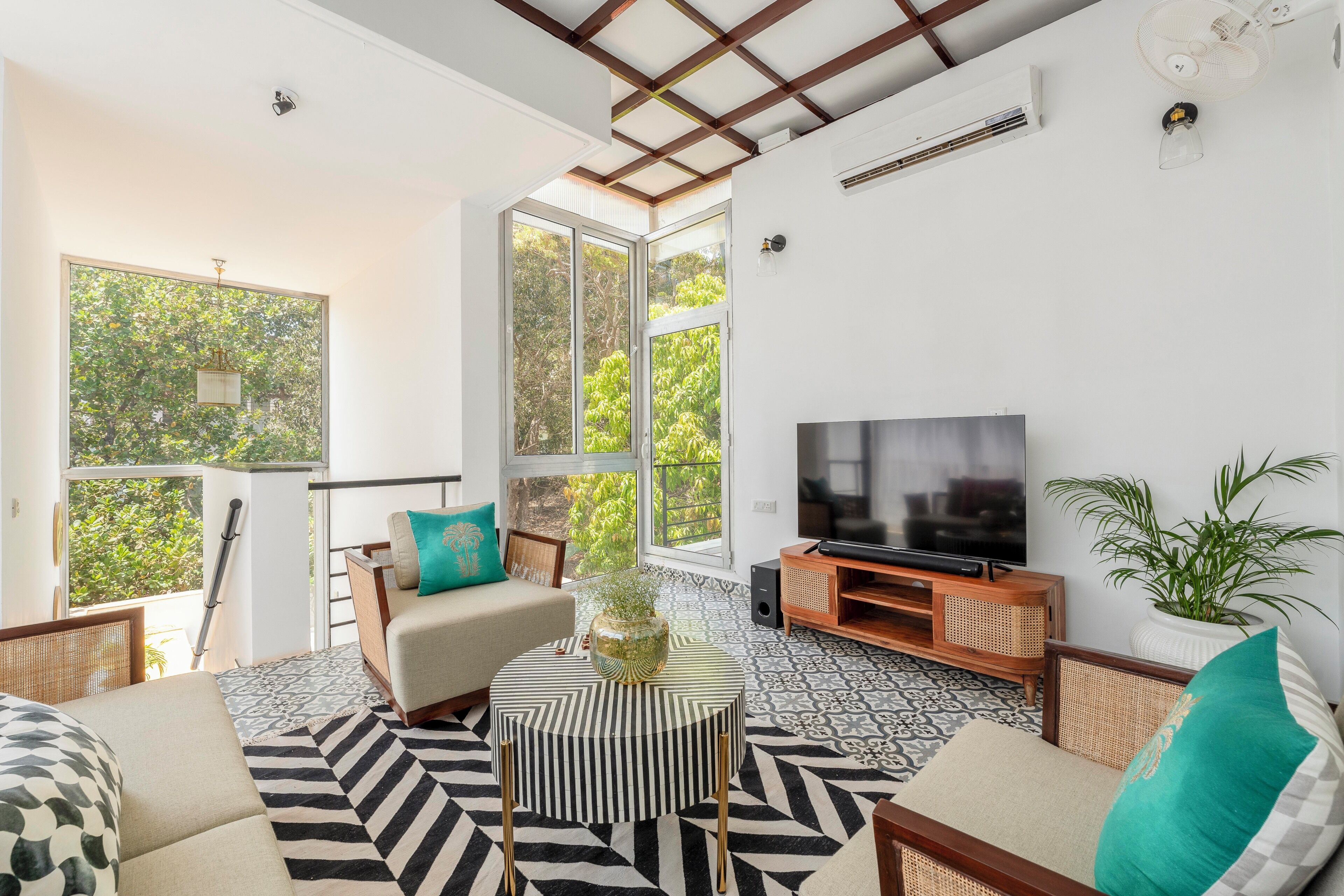 Ein Wohnzimmer mit Fernseher, Sofa und Couchtisch, großen Fenstern mit Blick auf Bäume und einem Deckenventilator.