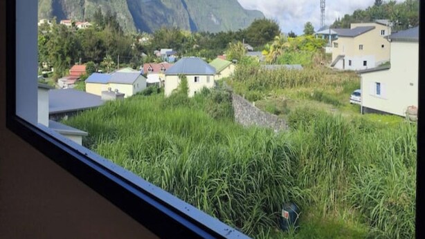 Une vue par la fenêtre sur un quartier résidentiel, avec des maisons, de la verdure et des montagnes en arrière-plan.
