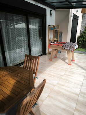 Mesa e cadeiras de madeira numa área externa coberta, com uma mesa de futebol e um armário com objetos.