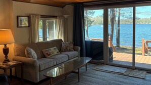 Un salon chaleureux avec un canapé, une table basse en verre et une lampe. Une grande fenêtre offre une vue sur un lac et une terrasse à l’extérieur.