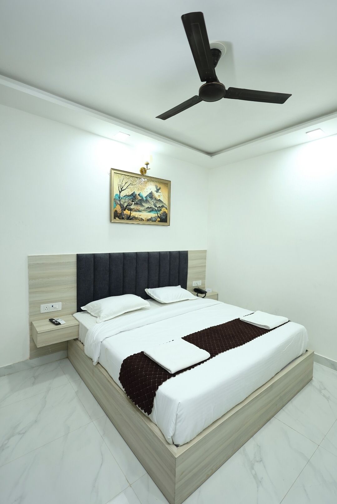 A bedroom with a bed, bedside tables, a ceiling fan, a wall-mounted painting, and a marble floor.