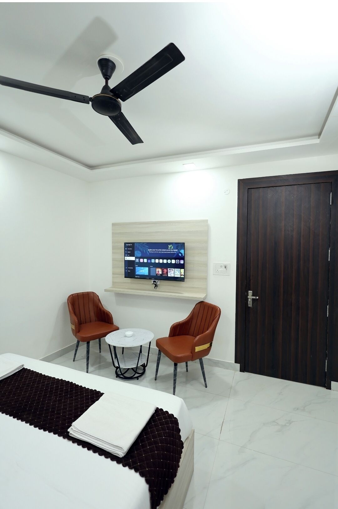 A hotel room with a bed, two armchairs, a small table, a ceiling fan, a mounted TV, and a wooden door.