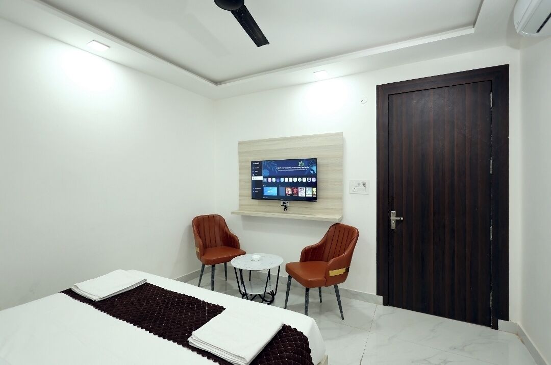 A hotel room with a bed, two chairs, a small table, a TV on the wall, and a dark wooden door.