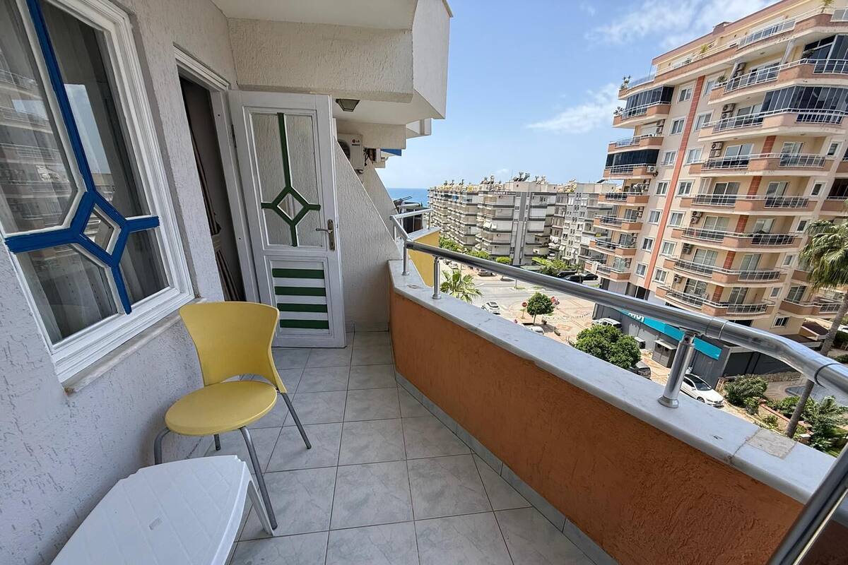 Un balcón con una silla amarilla, una mesa blanca y vistas de un edificio de varios pisos con balcones.