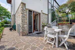A stone patio with a white table and chairs, a sliding glass door, and a small garden area with potted plants.