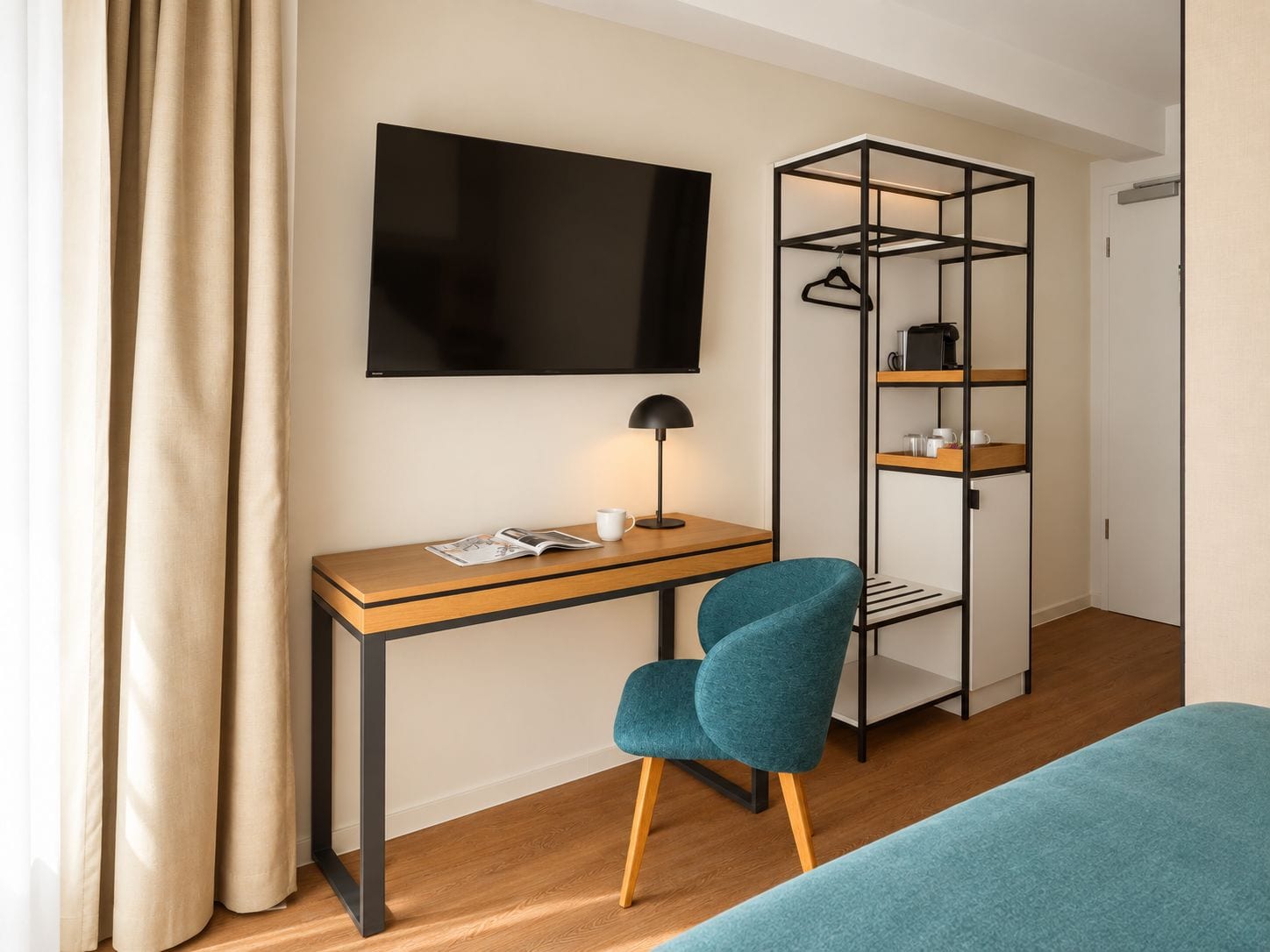 A modern hotel room with a wooden desk, a teal chair, a black TV, and a black metal wardrobe with shelves.