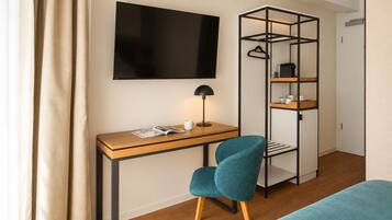 A modern hotel room with a wooden desk, a teal chair, a black TV, and a black metal wardrobe with shelves.