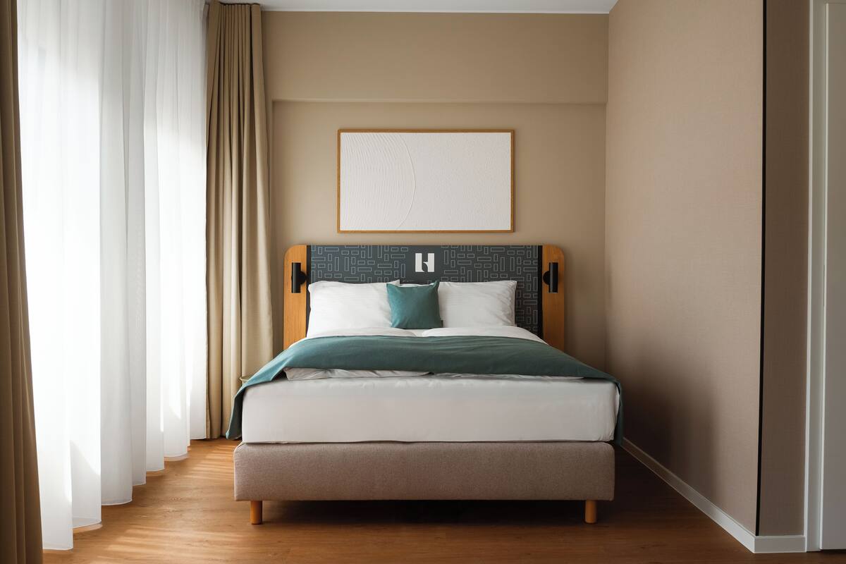 Una habitación de hotel con cama, cortinas, un cuadro en la pared y piso de madera.
