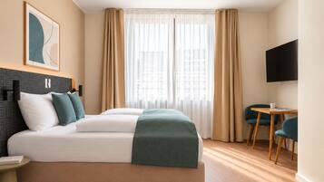 Habitación de hotel con cama, escritorio, silla, televisor y ventana con cortinas.