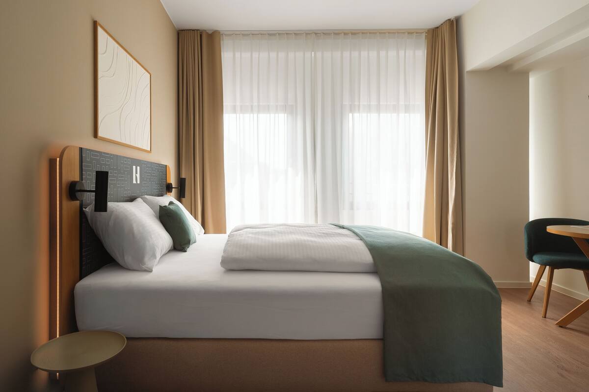 Una habitación de hotel con cama, mesita de noche, una silla y ventana con cortinas.