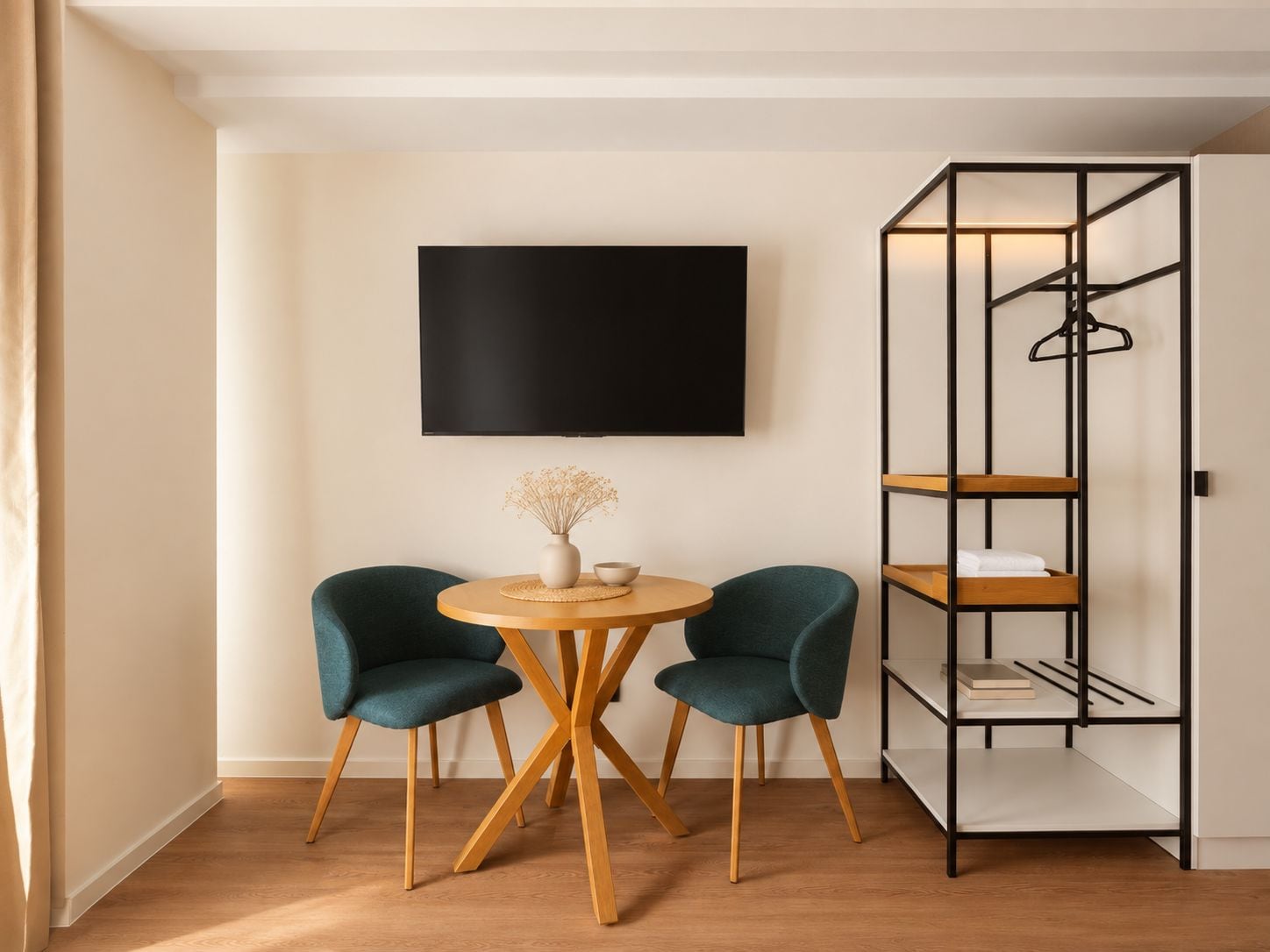 A modern room with a round wooden table, two teal chairs, a black TV on the wall, and a black metal shelving unit with a hanging rack.