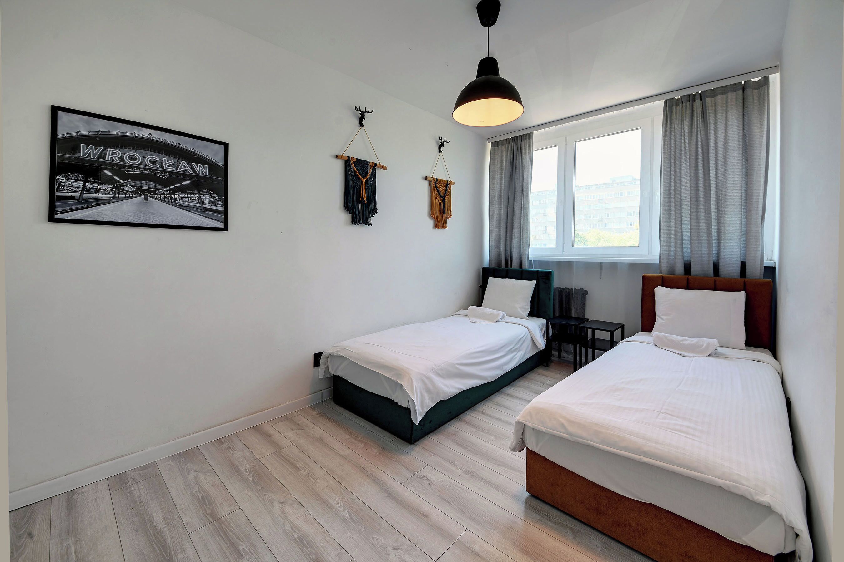 Quarto com duas camas, janela com cortinas, luminária pendente e quadro na parede.