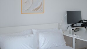 Una cama blanca con almohadas, un escritorio con monitor de computadora y un altavoz.