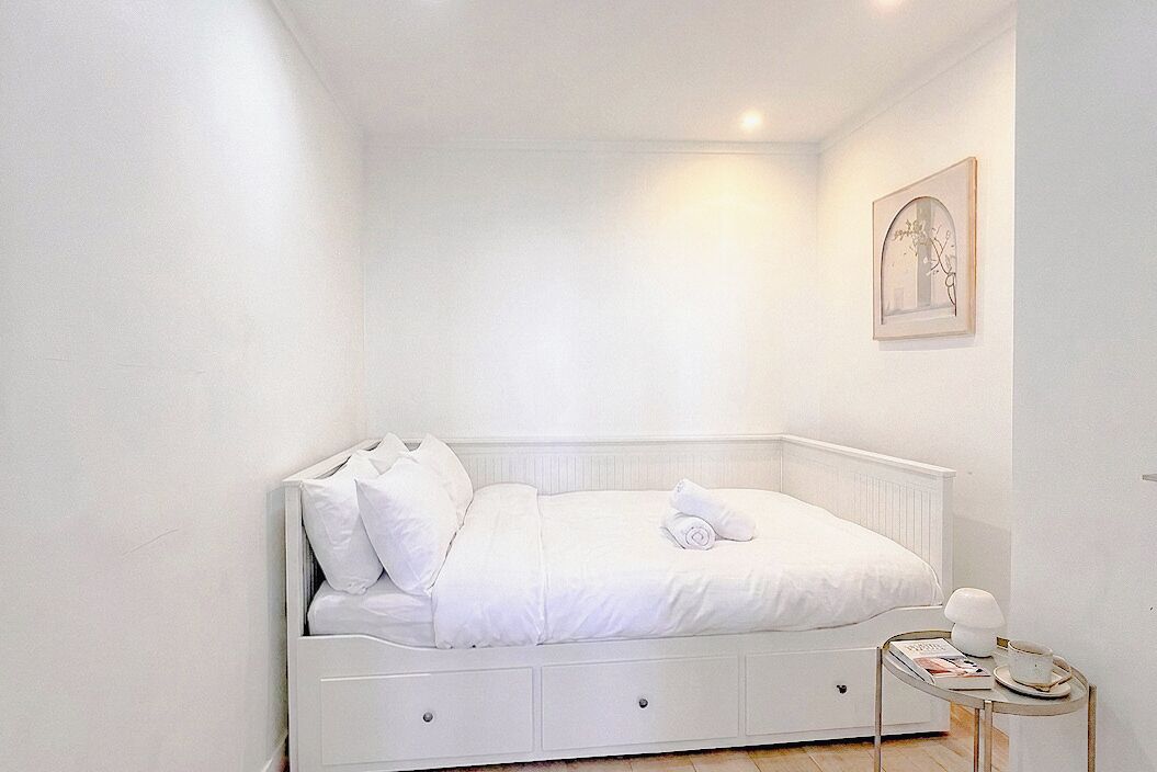 A white, single-bed room with a bedside table, a framed picture on the wall, and a small table with a lamp.