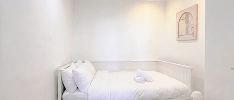 A white, single-bed room with a bedside table, a framed picture on the wall, and a small table with a lamp.