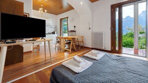 A modern room with a bed, a TV, a dining area, and a view of mountains. - Apartment 'Bilocale Ulrica' with Private Terrace and Wi-Fi (Nago–Torbole)