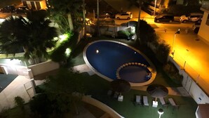 En swimmingpool med et nedsænket siddeområde, omgivet af liggestole og parasoller, med udsigt til parkerede biler og en bygning i baggrunden.