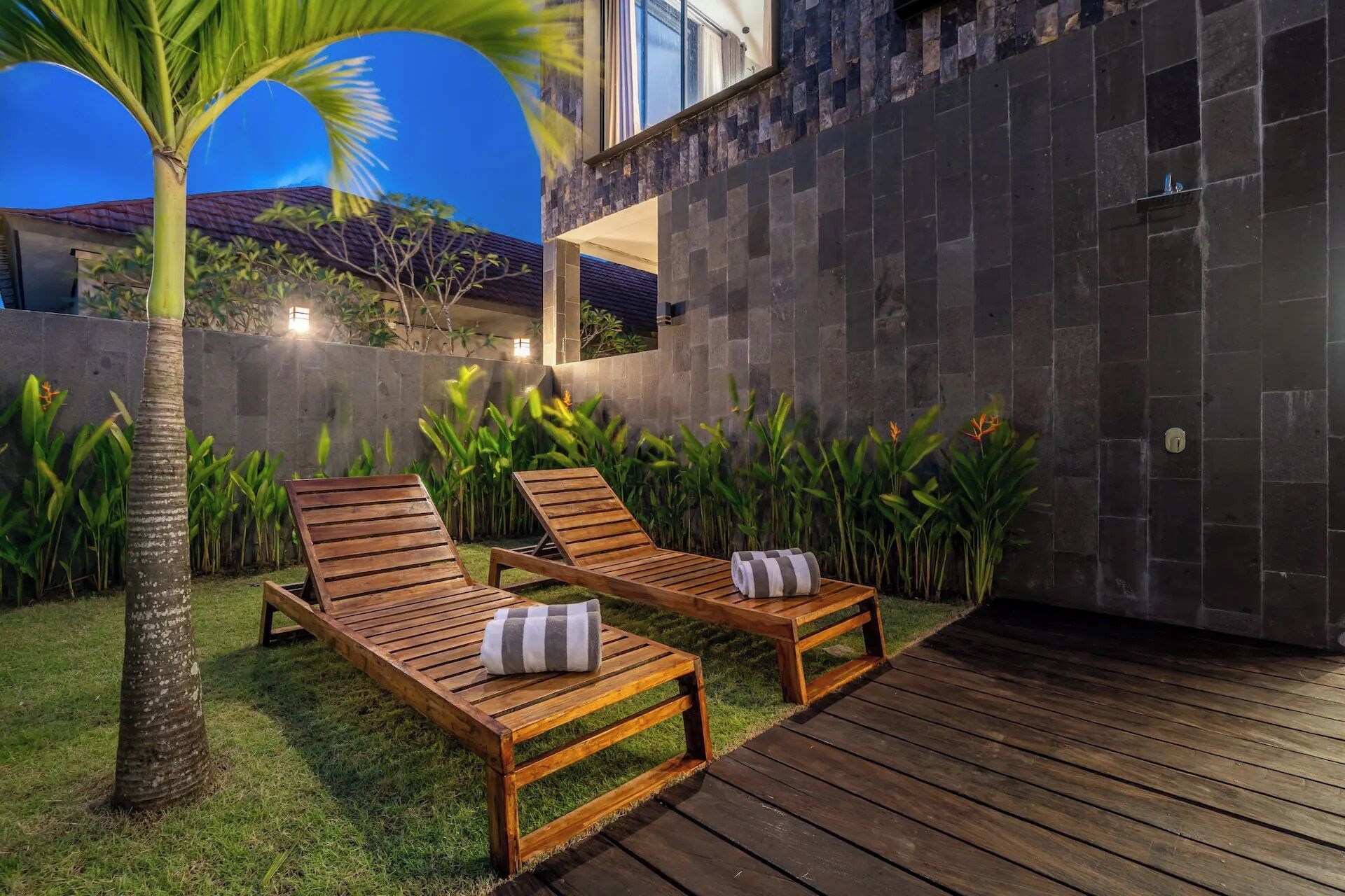 A patio with two wooden lounge chairs and a palm tree, surrounded by greenery and a stone wall.
