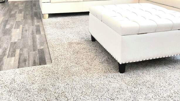 Una sala de estar moderna con un sofá seccional blanco, un pequeño reposapiés y una cocina al fondo.