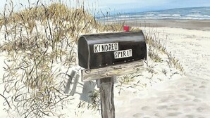 Un buzón con la etiqueta 'Kindred Spirit' sobre una playa arenosa, con césped y vista al océano.