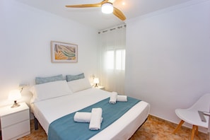 Ein Hotelzimmer mit Bett, Nachttischen, Deckenventilator, Fenster mit Vorhängen und Wandbild.