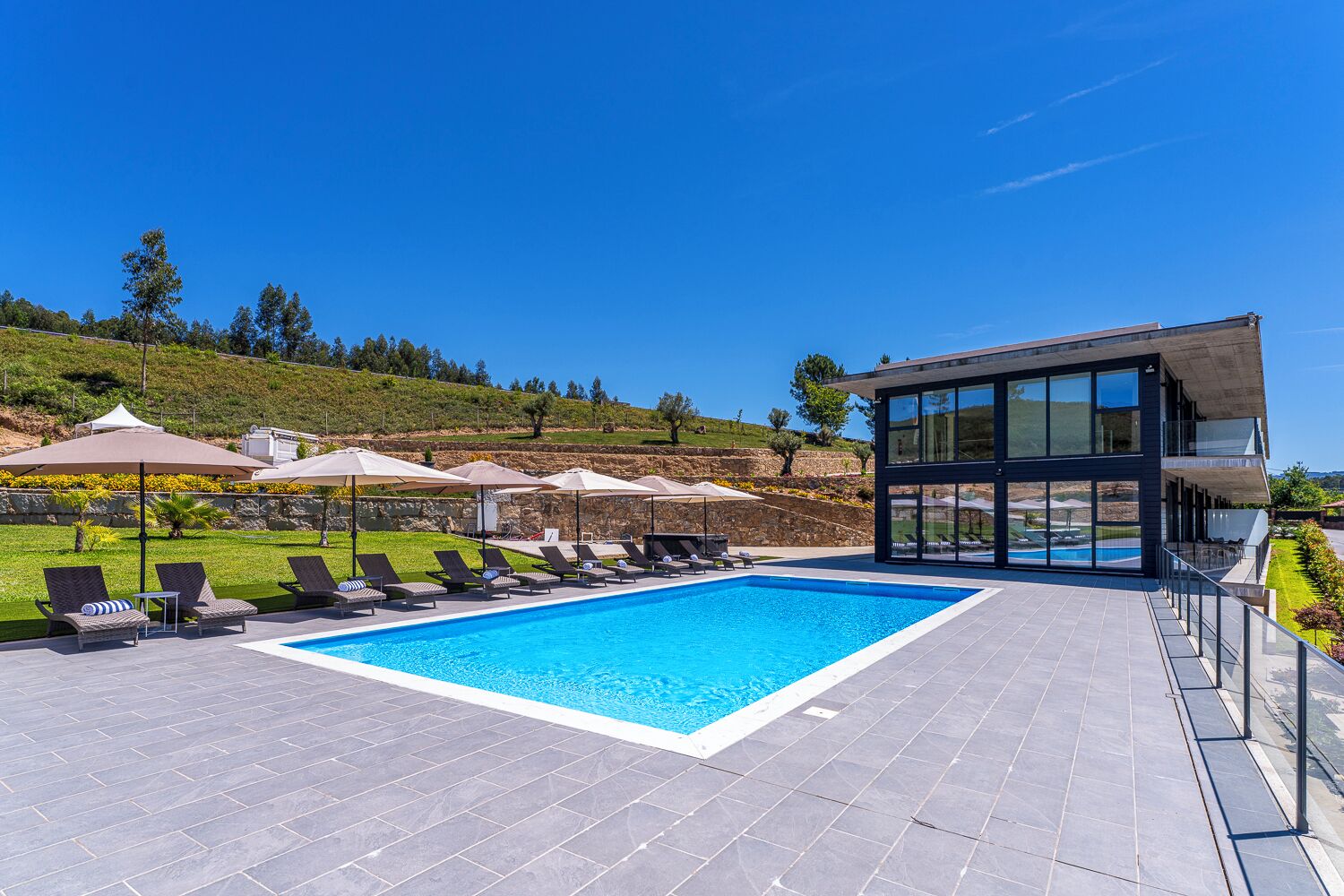 Un área de piscina moderna con sillas de descanso y sombrillas, una piscina azul cristalina y vistas a una colina.