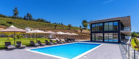 Un área de piscina moderna con sillas de descanso y sombrillas, una piscina azul cristalina y vistas a una colina.