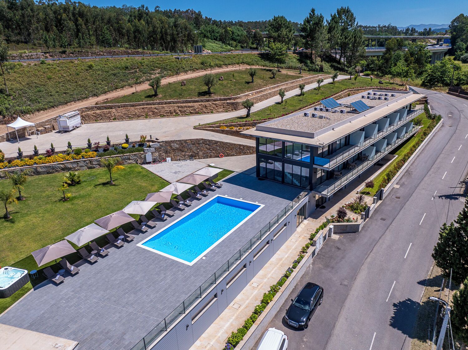 Un bâtiment moderne doté d’une piscine sur le toit, d’un espace extérieur couvert avec des chaises longues, et d’une route visible au loin.