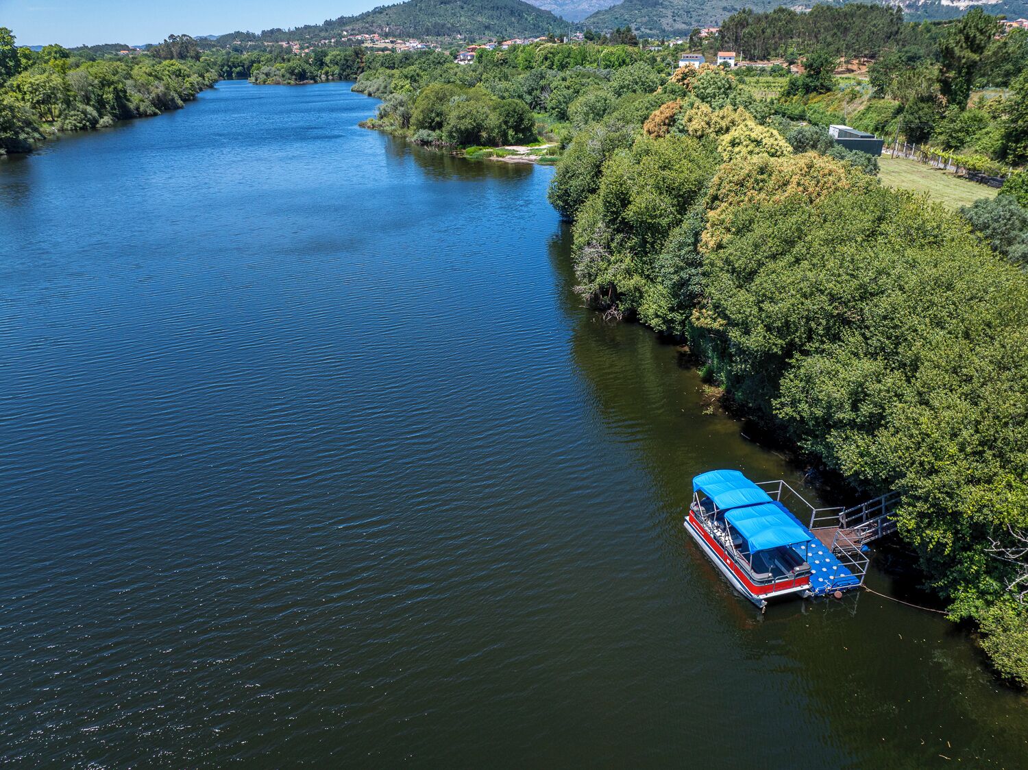 A river with a boat docked on the right bank, surrounded by dense greenery and a few buildings in the background.