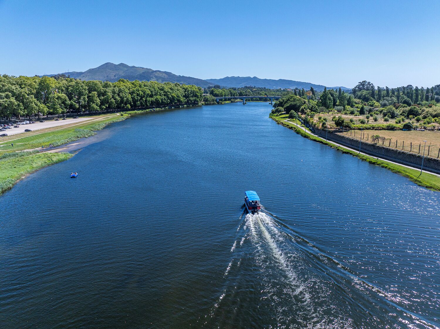 A river with a boat creating a wake, surrounded by greenery and a bridge in the distance.