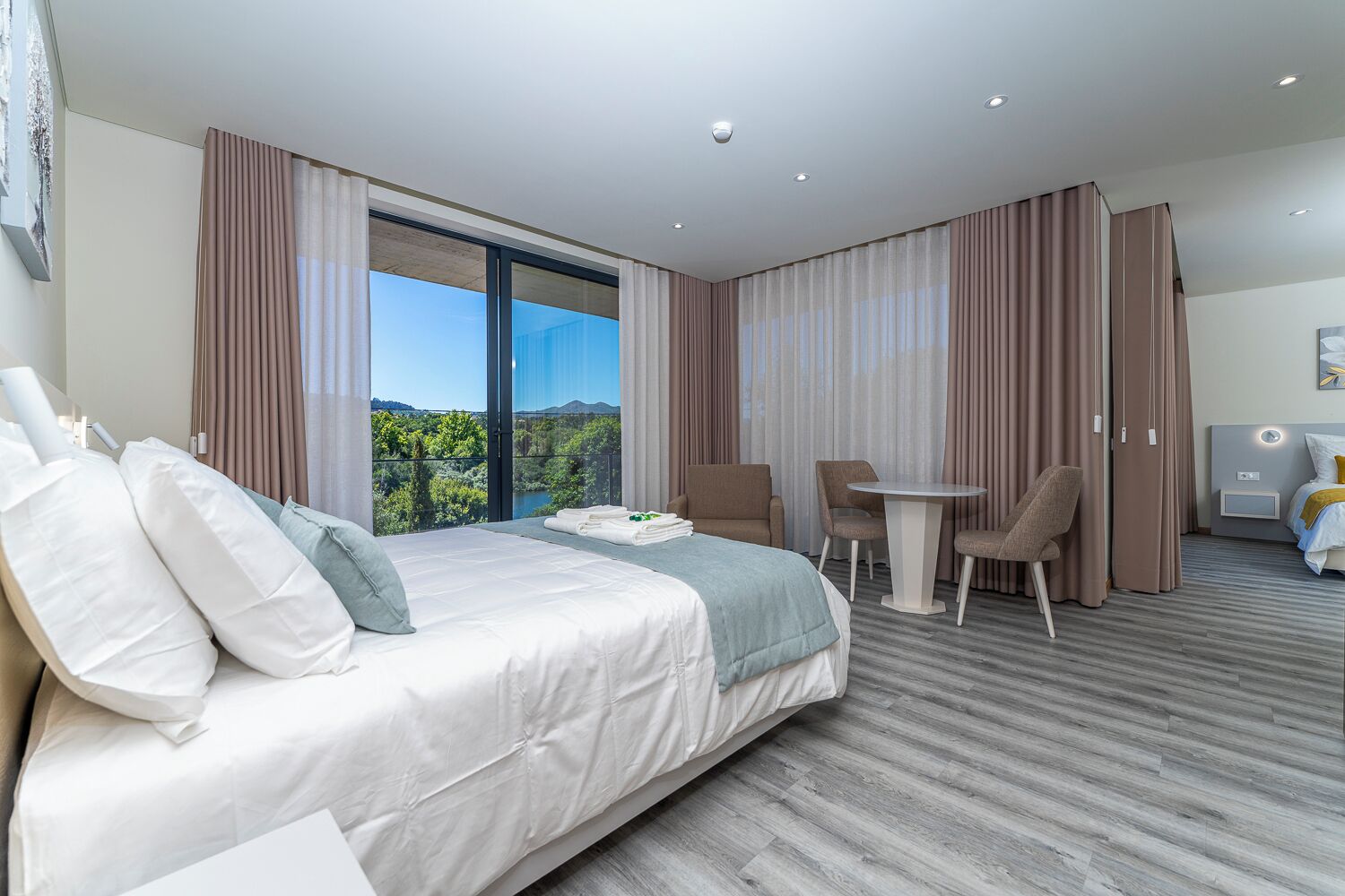 A hotel room with a bed, a small table, two chairs, a view of greenery, and a balcony.