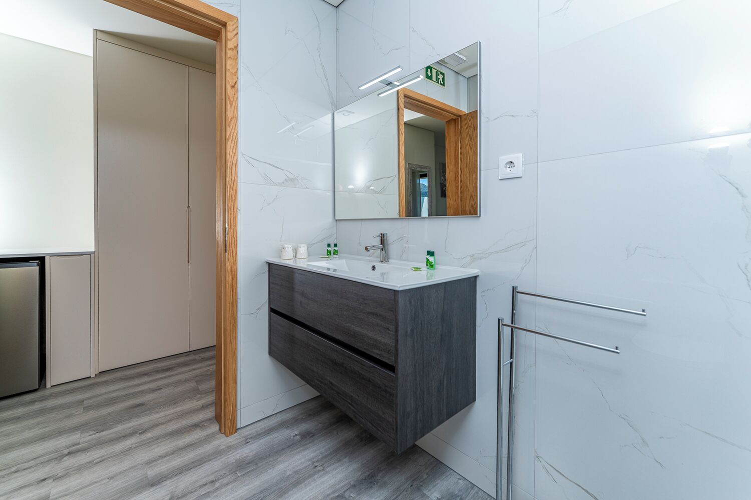 A modern bathroom with a marble wall, a wooden door, a dark wood vanity, and a glass shower enclosure.
