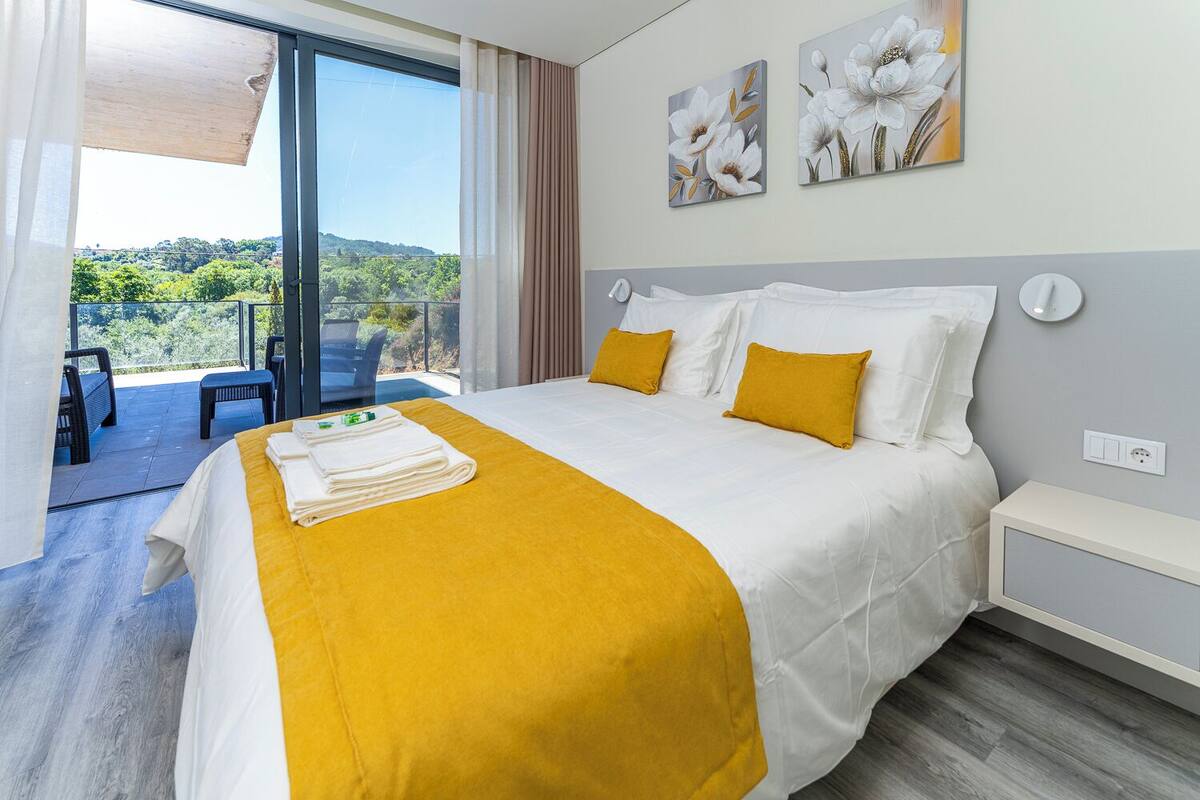 A hotel room with a bed, a balcony with chairs, and a view of greenery.