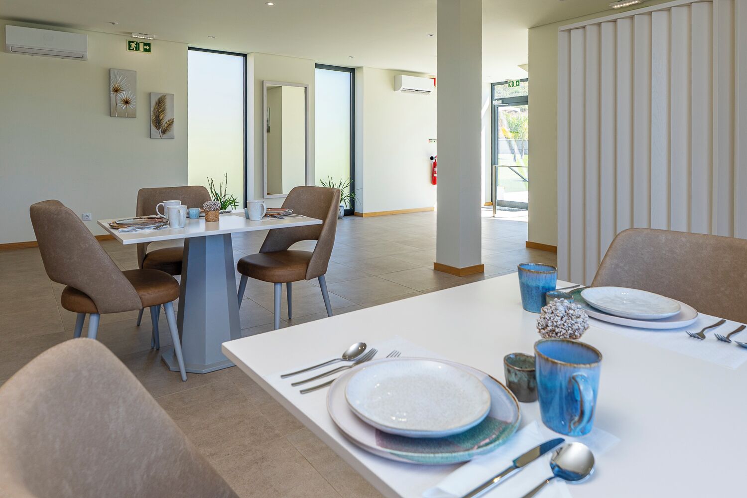 A dining area with a white table set for four, surrounded by chairs, with a view of the outdoors through large windows.