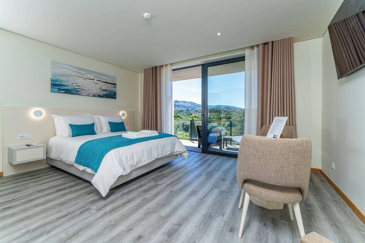 A hotel room with a bed, a chair, a nightstand, a painting, and a view of the outdoors.