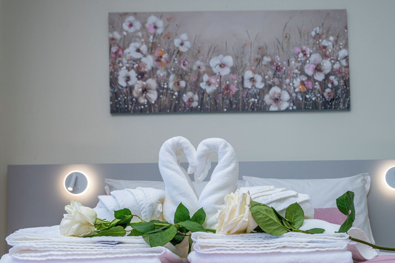 Un lit bien fait, avec des serviettes disposées en forme de cœur, une rose et des feuilles vertes sur le lit, et un tableau floral accroché au mur derrière.